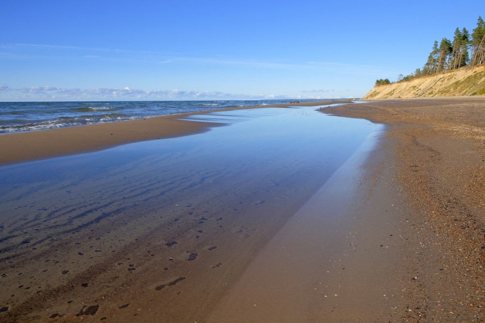 Klusas jūras piekraste ar smilšu pludmali un stāvkrastu zem zilām debesīm
