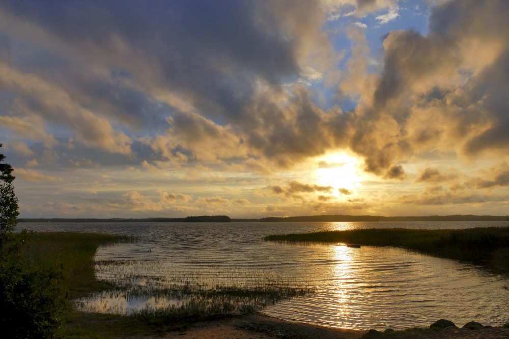 Saulriets virs ezera ar zeltainu atspīdumu un dramatiskām mākoņu formām
