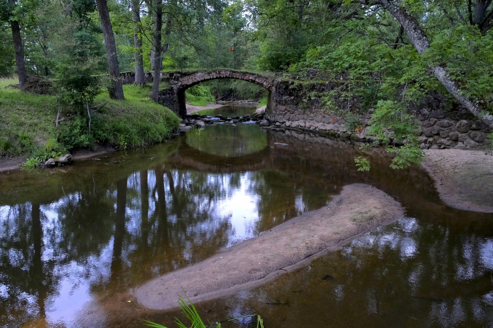 Vēsturisks mūra tilts meža ainavā ar ūdens atspulgiem