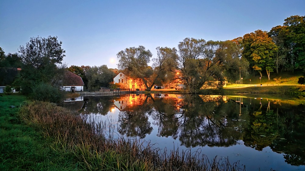 Calm pond view in Aizpute with tree and building reflections at dusk