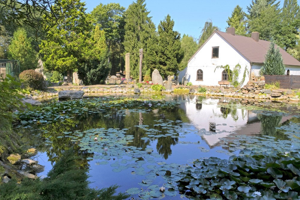 Dīķis ar ūdensrozēm un baltu ēku zaļā dārza ainavā