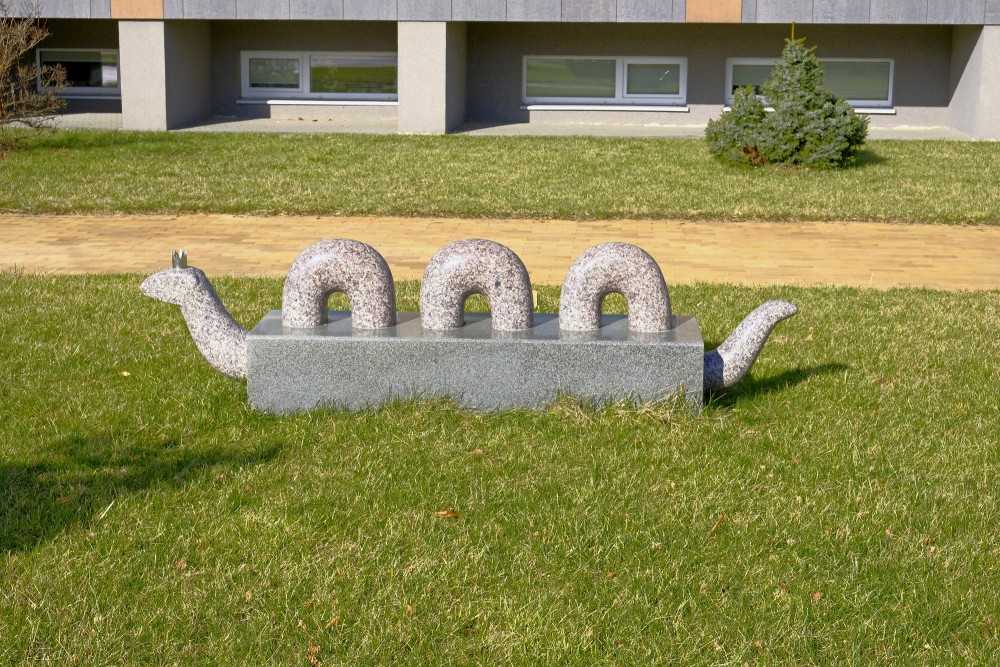 Akmens skulptūra čūskas tēlā uz zāliena pie celiņa