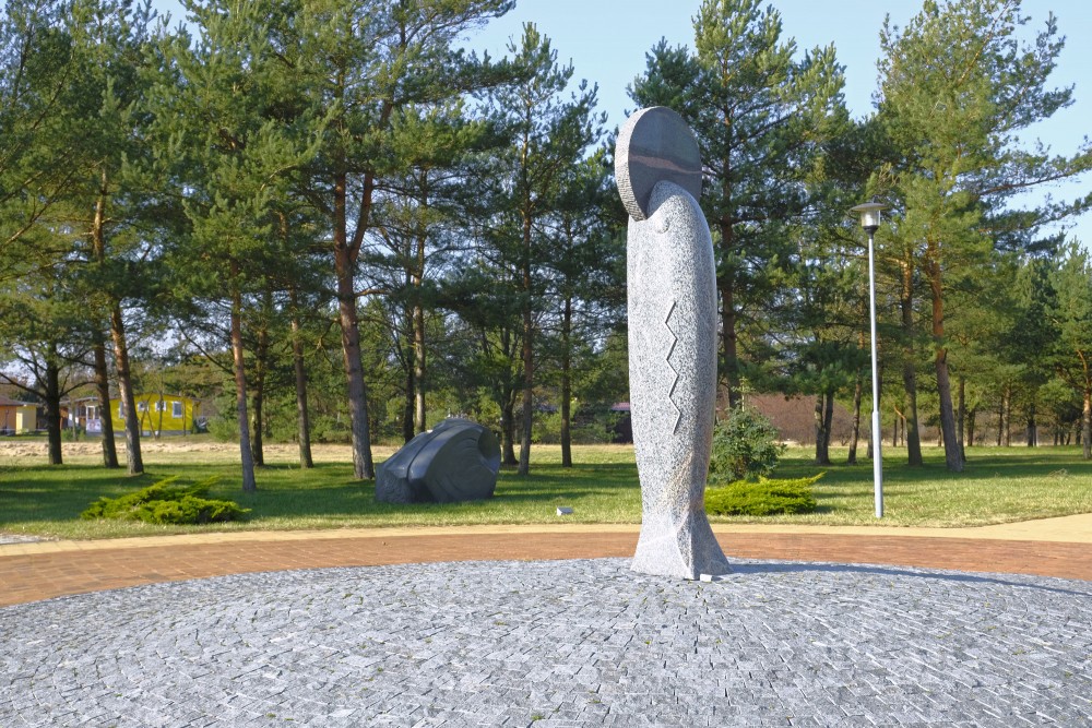 Vertikāla akmens skulptūra parkā ar priedēm un bruģētu laukumu