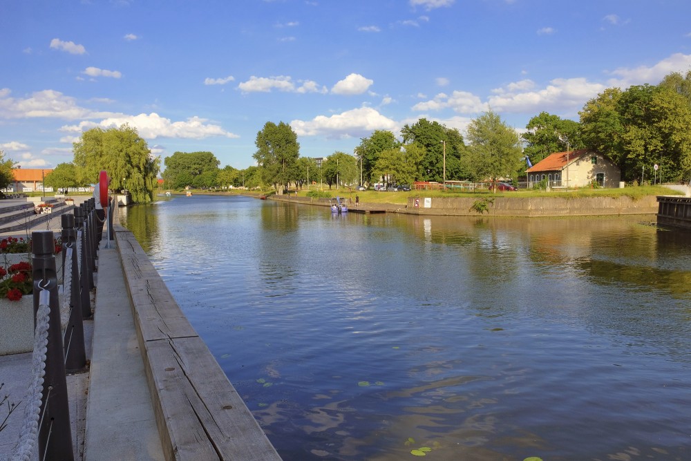 Danes upes skats no promenādes ar koka krastmalu un koku ieskautiem krastiem